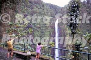 A 1970s colour photo of Akaka Falls in Hawaii, featuring a high waterfall plunging into a green gorge with tourists in 1970s clothing standing at a railing.