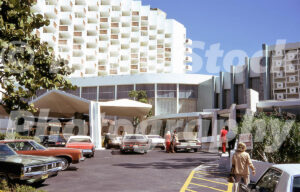 A 1970s color photo of the Americana Hotel exterior in Bal Harbour, Florida, featuring its tiered white balconies, a modern geometric porte-cochère, and a parking lot with mid-seventies American cars.