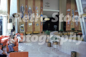 A 1970s color photo of the Americana Hotel lobby in Bal Harbour, Florida, featuring massive wall mosaics of historical figures, orange lounge chairs, and the entrance to the Bal Masque supper club.