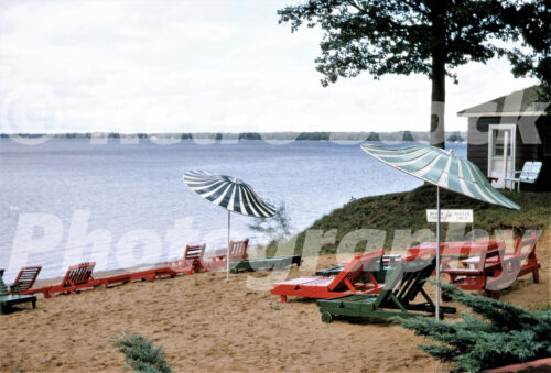 A 1960s color photo of the sandy beach at Johnson’s Rustic Resort on Houghton Lake in Prudenville, Michigan, featuring red and green wooden lounge chairs and striped umbrellas.