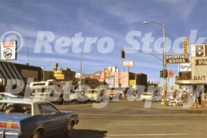 A 1980s color photo of a busy street corner in Bishop, California, featuring Levi's The Toggery, Sierra Liquor, and Joseph's Bi-Rite Market with vintage 1970s cars in the foreground.
