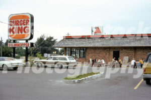 A 1970s color photo of a Burger King restaurant in Marathon, Florida, featuring a classic wood-paneled station wagon and a white Ford Thunderbird in the parking lot, with people eating at outdoor picnic tables.