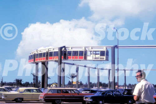 A 1960s color photo of the suspended monorail at Busch Gardens in Tampa, Florida, featuring red and white passenger cars traveling over a parking lot filled with mid-sixties American station wagons and sedans.