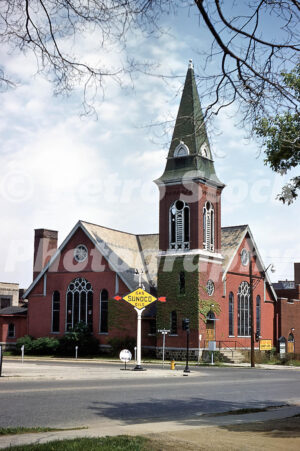 A 1950s color photo of the red brick Central Methodist Church in Flint, Michigan, featuring a tall ivy-covered spire and a vintage yellow Sunoco Gas Oils sign on the corner.
