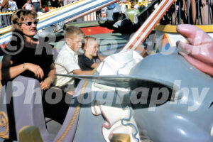 A 1960s color photo of a woman in cat-eye sunglasses and two young boys riding the Dumbo the Flying Elephant attraction at Disneyland in California.