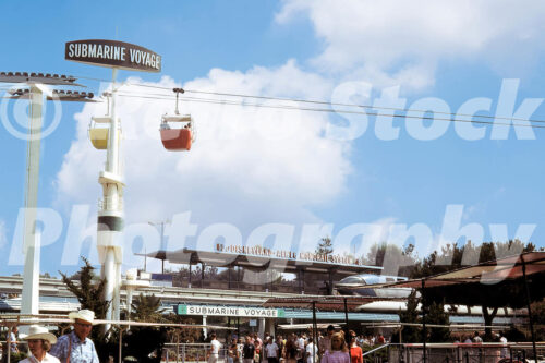 A 1960s color photo of the Tomorrowland area at Disneyland in California, featuring the sleek silver Monorail, the Submarine Voyage entrance, and Skyway gondolas passing overhead against a bright blue sky.