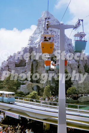 A 1970s color photo of the Matterhorn Bobsleds at Disneyland in California, showing orange and blue Skyway gondolas passing in front of the snow-capped mountain and over the Monorail track.