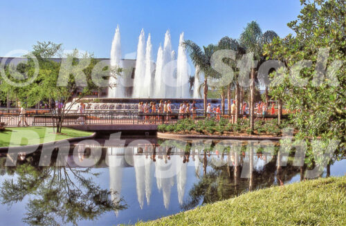 A 1980s color photo of the Fountain of Nations at Epcot Center in Florida, showing large water plumes reflecting in a calm pond with guests walking across a bridge and palm trees in the foreground.