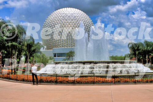 A 1980s color photo of the Fountain of Nations at Epcot Center in Florida, featuring large white water plumes and the Spaceship Earth geosphere under a cloudy blue sky.