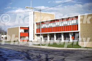 A 1960s color photo of the Flint Fire Department Headquarters in Michigan, featuring mid-century modern architecture with red panels, glass bay doors, and tan brickwork.