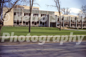 A 1960s color photo of the Flint Public Library in Michigan, featuring mid-century modern architecture with grey panels, floor-to-ceiling windows, and a manicured green lawn.