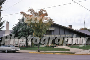 A 1970s color photo of the Frankenmuth State Bank in Michigan, featuring a long, low-profile building with a grey timber-framed upper story, white brick, and autumn trees.