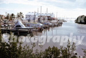 A 1970s color photo of Gilbert's Resort & Marina in Key Largo, Florida, featuring several yachts and motorboats docked in the calm canal, with the main restaurant and "Boat & Motor Sales" building in the background.