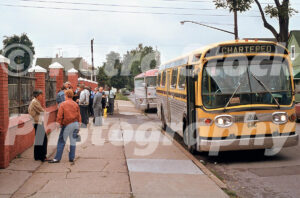A 1980s colour photo of a yellow chartered GM bus parked on the street with people gathering near the brick wall of the Holy Cross Hungarian Church in 1987.