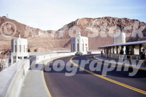 A 1970s colour photo of the Art Deco intake towers and a classic car driving across the crest of Hoover Dam in 1970s.