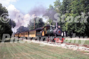 A 1980s color photo of Huckleberry Railroad Steam Locomotive No. 2 pulling a string of vintage yellow passenger cars through a wooded area in Flint, Michigan.