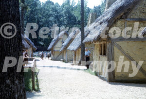 A 1960s color photo of Jamestown Festival Park in Virginia, featuring a row of wattle-and-daub houses with thatched roofs and guests walking along a dirt path through the historical recreation.
