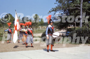 A 1960s color photo of historical interpreters at Jamestown Festival Park in Virginia, featuring men in seventeenth-century armor and red-plumed helmets, including a drummer and a flag bearer holding the Cross of St. George.