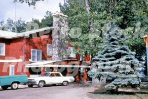 A 1960s color photo of the main lodge at Johnson’s Rustic Resort in Prudenville, Michigan, featuring red wood siding, a large fieldstone chimney, and a cream-colored vintage sedan.