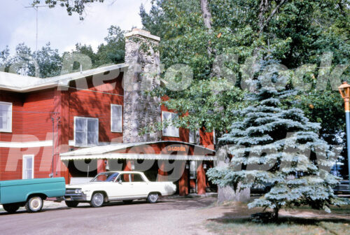 A 1960s color photo of the main lodge at Johnson’s Rustic Resort in Prudenville, Michigan, featuring red wood siding, a large fieldstone chimney, and a cream-colored vintage sedan.
