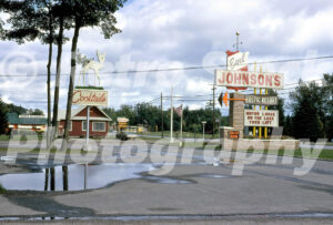 A 1960s color photo of the entrance to Johnson’s Rustic Resort in Michigan, featuring a large stone pylon sign for Earl Johnson’s and a "Cocktails" sign topped with a white deer statue.