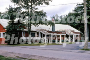 A 1960s color photo of the main restaurant building at Johnson’s Rustic Resort in Prudenville, Michigan, featuring red wood siding, white-trimmed porches, and a large decorative candle on the roof.