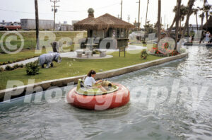 A 1980s color photo of a girl operating a red and green bumper boat at Jungleland in North Carolina, with a thatched-roof tiki hut, a small elephant statue, and a vintage car on a nearby road.