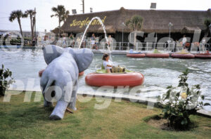 A 1980s color photo of a young girl in a red bumper boat at Jungleland in North Carolina, with a large elephant statue spraying water and the thatched-roof main building in the background.
