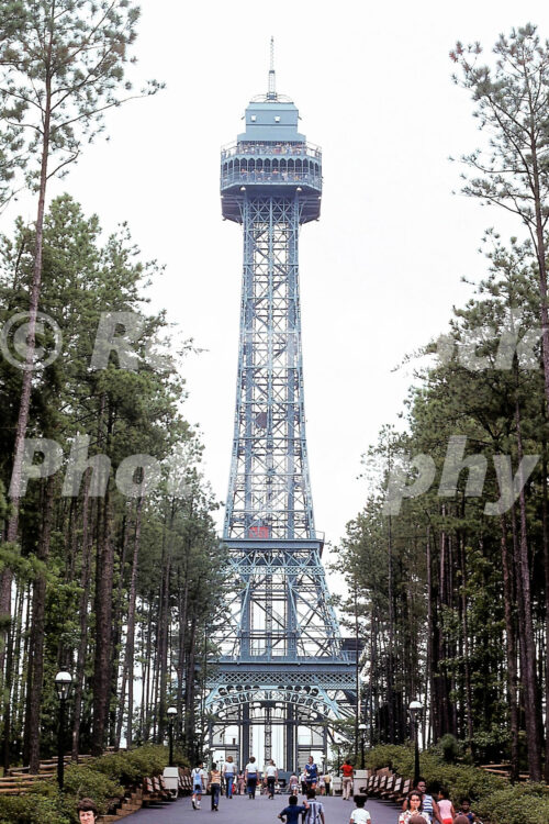 A 1970s color photo of the Eiffel Tower at Kings Dominion in Virginia, showing the blue steel lattice structure rising above a wooded pedestrian path lined with benches and globe street lamps.