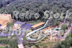 A 1970s color photo of the Shenandoah Lumber Company log flume at Kings Dominion in Virginia, featuring a log boat carrying passengers down a steep turquoise chute into a splashdown pool surrounded by pine trees.