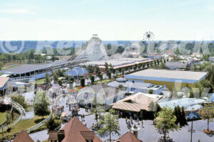 A 1970s color photo of the Coney Island area at Kings Island in Ohio, featuring The Racer wooden roller coaster, a Ferris wheel, and various game buildings under a clear sky.