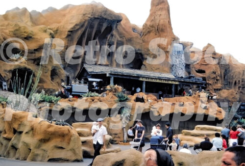 A 1960s color photo of the Calico Mine Ride at Knott's Berry Farm in California, featuring the rocky mountain exterior, a waterfall, and the Calico Mine Co. station with guests in the foreground.