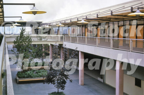 A 1960s color photo of the second-level pedestrian walkways at Lloyd Center in Portland, Oregon, featuring glass-walled offices, mesh railings, and yellow disk-shaped pendant lights.
