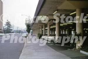 A 1960s color photo of the "M&F Customer Pick-Up" lane at Lloyd Center in Portland, Oregon, featuring a long row of concrete pillars, parked vintage cars, and Manning's Coffee Cafe.
