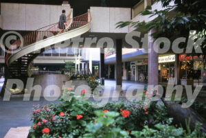 A 1960s color photo of a curved spiral staircase with orange railings in an outdoor garden court at Lloyd Center in Portland, Oregon, featuring shops like Columbian Optical and National Clothing.