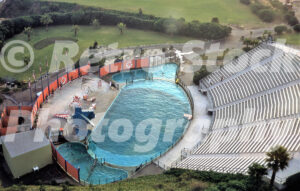 A 1960s colour photo of Marineland of the Pacific in California, featuring an aerial view of the outdoor stadium, a large turquoise performance pool, and tiered concrete seating overlooking the Pacific coast.