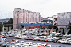 A 1970s color photo of a blue monorail train traveling above a crowded parking lot filled with vintage cars at Hersheypark in Pennsylvania, with large industrial grain silos in the background.