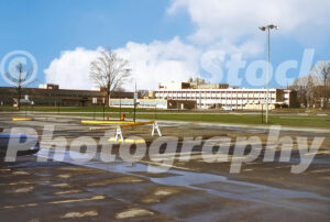 A 1960s color photo of the Mott Community College campus in Flint, Michigan, featuring mid-century academic buildings across a large asphalt parking lot with puddles.