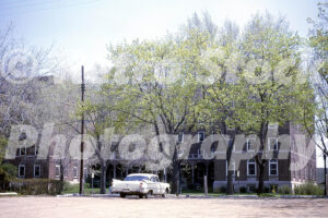 A 1960s color photo of the brick Nurses Residence building at Hurley Hospital in Flint, Michigan, partially obscured by budding spring trees with a white 1959 Dodge parked in front.