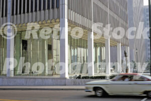 A 1960s color photo of a white car driving past the glass-walled lobby of the One Woodward Avenue building in Detroit, Michigan in 1967.
