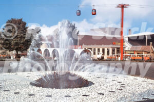 A 1960s color photo of the International Fountain at the Seattle World's Fair in Washington, featuring large water jets spraying from a spiked dome, with the Skyride gondolas and fair pavilions in the background.
