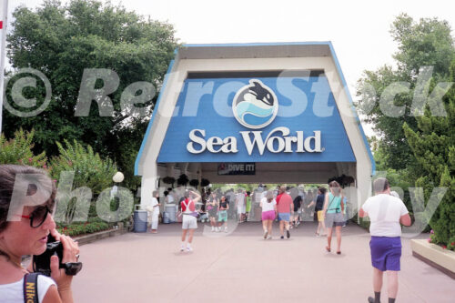 A 1990s color photo of the main entrance to Sea World in Orlando, Florida, featuring a large blue A-frame sign with the classic orca logo and guests walking toward the gates.
