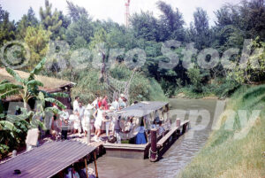 A 1960s color photo of La Salle’s River Adventure at Six Flags Over Texas in Arlington, featuring guests boarding a flat-bottomed boat from a wooden dock surrounded by tropical foliage.