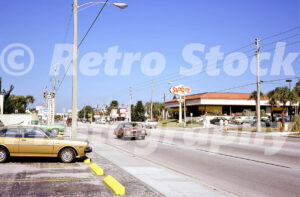 A 1980s color photo of Gulf Boulevard in St. Petersburg, Florida, featuring a Sambo's Restaurant with its iconic oval sign, a gold Toyota Corolla, and a brown 1978 Buick Regal driving on the sunny roadway.