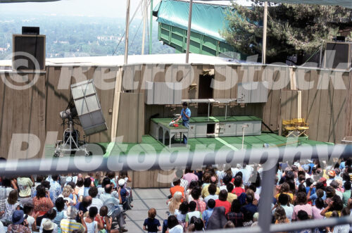A 1970s color photo of a special effects demonstration stage at Universal Studios Hollywood, featuring a staff member on a green-carpeted set in front of a seated audience and a large light reflector.