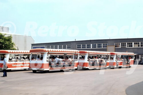 A 1970s color photo of several orange and white Universal Studios Glamor Trams parked in a staging area in front of a grey studio building in Hollywood, California.