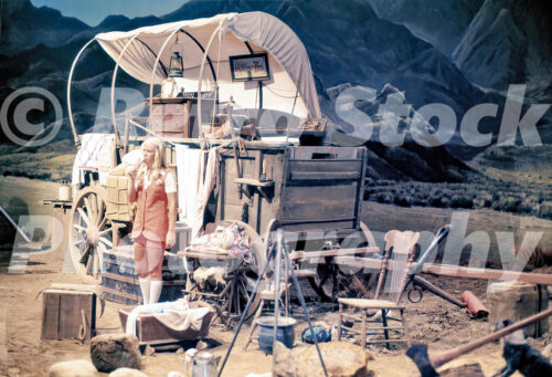 A 1970s color photo of a female tour guide in an orange uniform standing in front of a highly detailed covered wagon set on the Universal Studios Hollywood backlot.