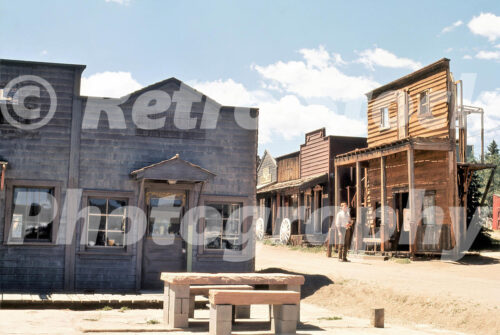 A 1970s color photo of the Western Street backlot at Universal Studios Hollywood in California, featuring weathered wooden storefronts, a dirt road, and a costumed actor standing near a porch.