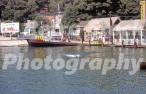 A 1970s color photo of the Jaws attraction at Universal Studios Hollywood in California, featuring the Orca boat at a dock in Amity Island with a shark fin visible in the water.