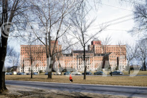 A 1950s color photo of a woman in a red coat walking past the large brick Veterans Hospital in Allen Park, Michigan in 1955.